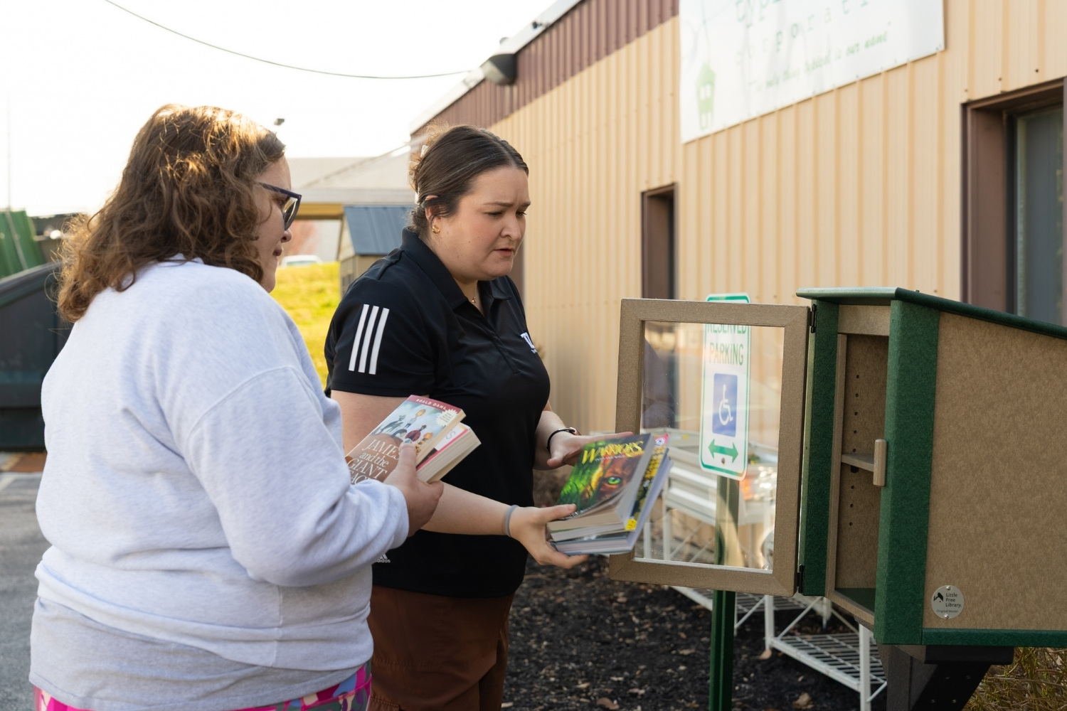 Two women place books into an open Little Free Library book box outside a building.