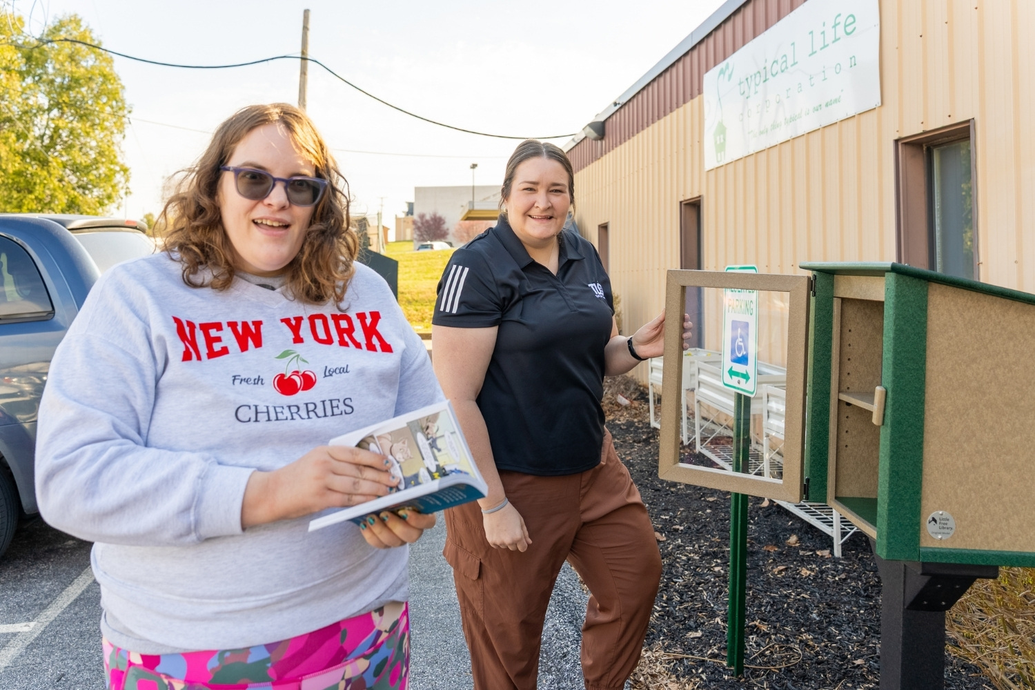 A woman in a “New York Cherries” sweatshirt and another woman in a black shirt stand beside an open Little Free Library book box, holding books and smiling outside a building.