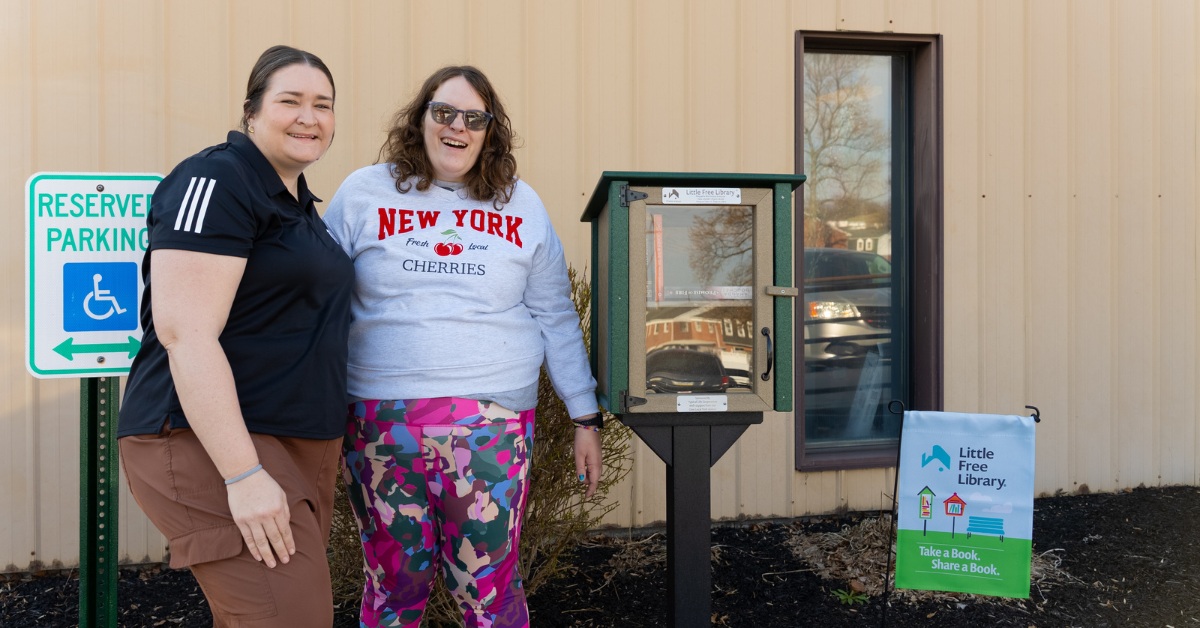 Two women stand and smile in front of a Little Free Library book box outside a building, next to a sign for accessible parking.