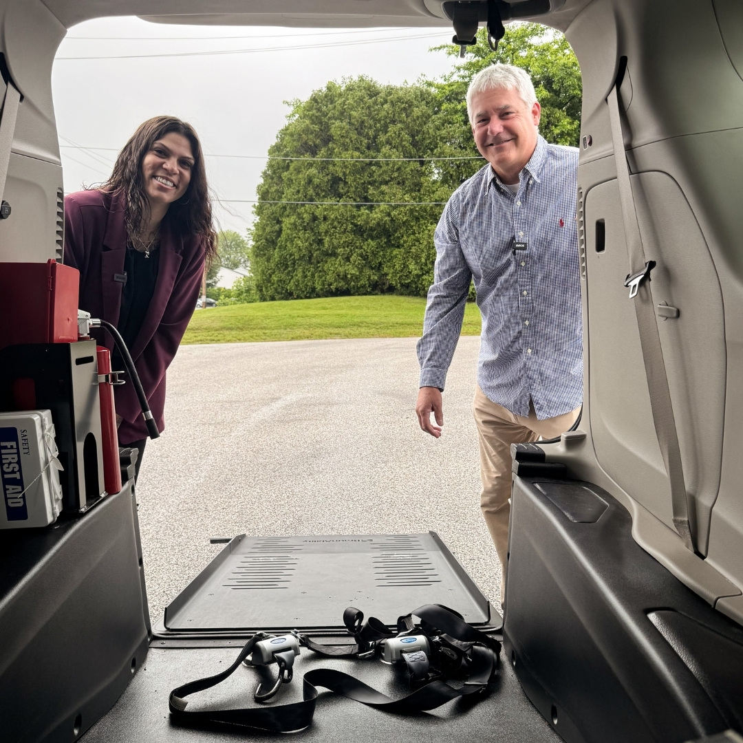 View from inside an accessible minivan looking outward, with a man and a woman smiling at the open ramp, securement straps visible in the foreground.