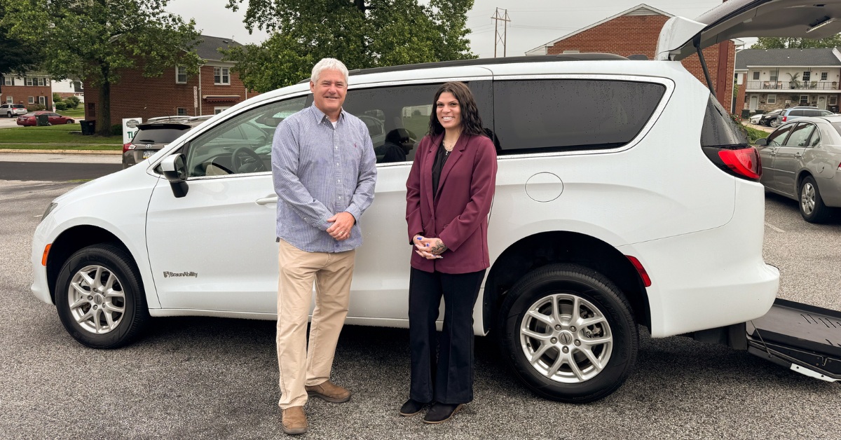 A man and a woman stand smiling in front of a white accessible minivan with a ramp, parked in a lot.